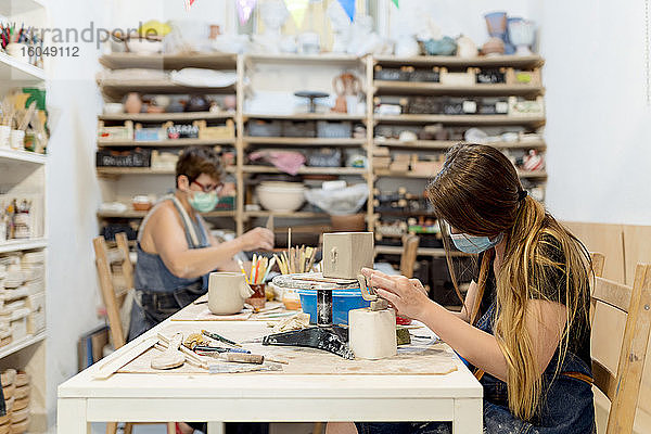 Frauen mit Masken bei der Keramikherstellung auf einer Bank in einer Werkstatt