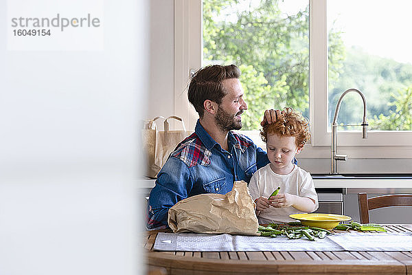 Lächelnder Mann sieht seinen Sohn an  der eine grüne Erbse hält  während er in der Küche sitzt