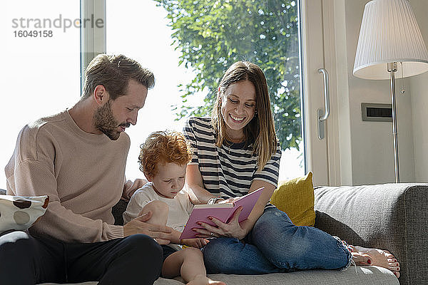 Lächelnde Frau  die ein Bilderbuch liest  während sie neben ihrem Sohn und einem Mann auf dem Sofa im heimischen Wohnzimmer sitzt