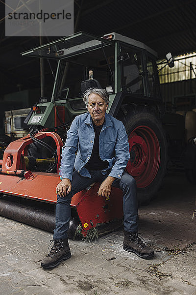Älterer Mann auf einem Bauernhof sitzt auf einem Traktor in einer Scheune