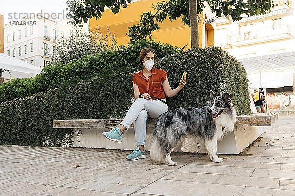 Junge Frau mit Schutzmaske sitzt auf einer Bank in der Stadt und fotografiert mit ihrem Smartphone