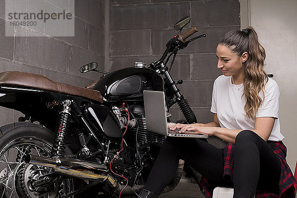 Junge Frau  die einen Laptop benutzt und neben einem Motorrad sitzt