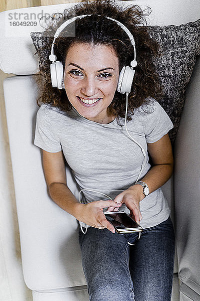 Porträt einer lächelnden jungen Frau  die mit Kopfhörern und Smartphone auf der Couch liegt und Musik hört