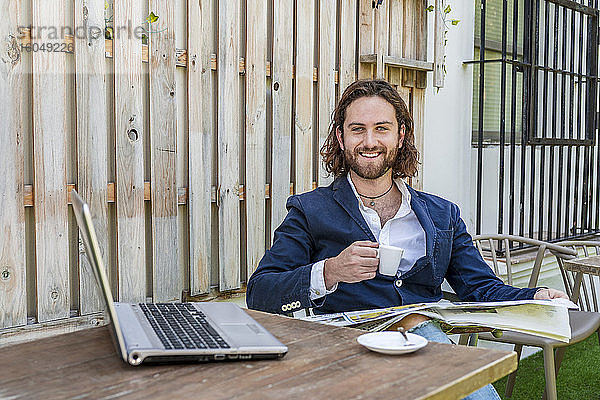 Lächelnder gutaussehender junger Geschäftsmann liest Zeitung und hält eine Kaffeetasse neben seinem Laptop am Tisch in einem Café