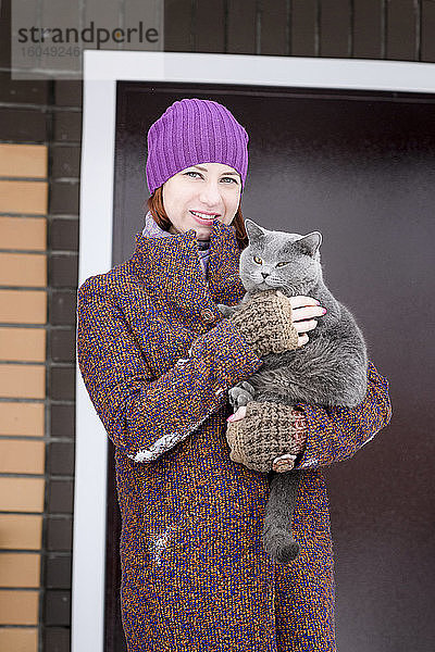 Schöne Frau in warmer Kleidung mit Britisch-Kurzhaar-Katze vor der Haustür