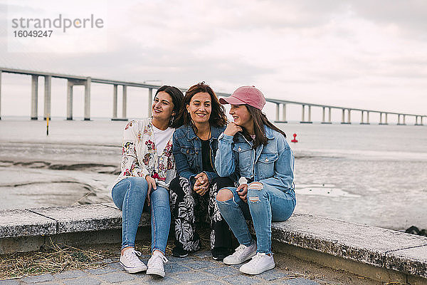 Lächelnde Mutter mit Töchtern auf einer Stützmauer am Meer sitzend