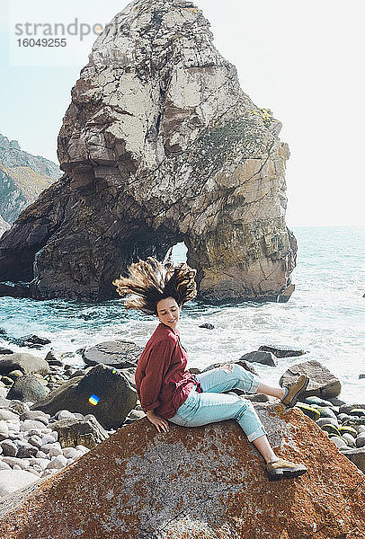 Unbekümmerte junge Frau schüttelt den Kopf  während sie auf einem Felsen am Strand von Ursa sitzt  Portugal