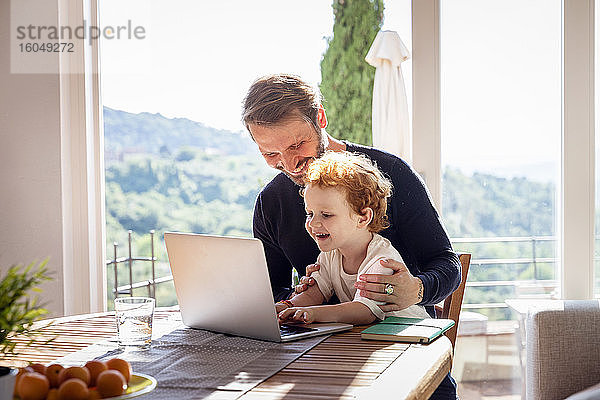 Lächelnder Mann sieht seinen Sohn an  der einen Laptop benutzt  während er im Wohnzimmer am Fenster sitzt