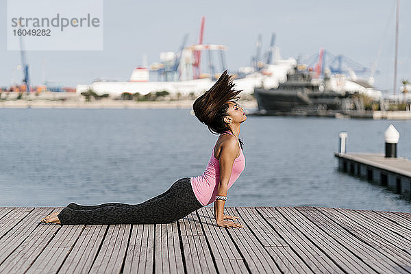 Weibliche Athletin mit geschlossenen Augen übt Kobra-Pose auf Pier gegen das Meer am Hafen