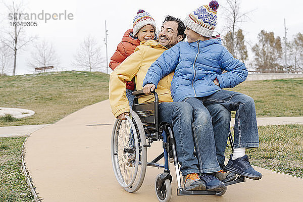 Glücklicher Mann mit verspielten Söhnen  die im Rollstuhl auf einem Fußweg im Park fahren