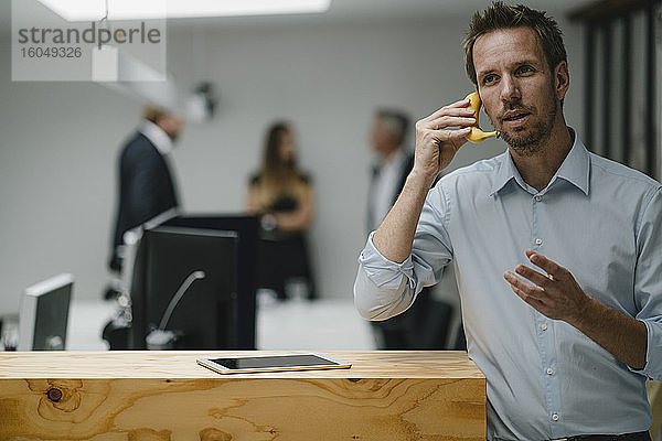 Geschäftsmann steht in offener Bürotür und benutzt eine Banane als Telefon