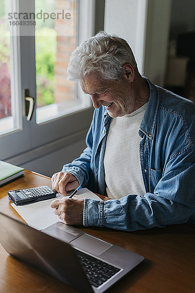 Glücklicher älterer Mann  der zu Hause mit Taschenrechner und Laptop auf Papier schreibt