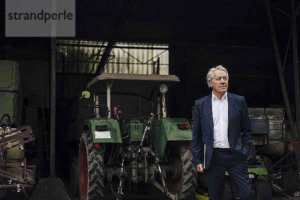 Älterer Geschäftsmann mit Laptop auf einem Bauernhof mit Traktor in der Scheune