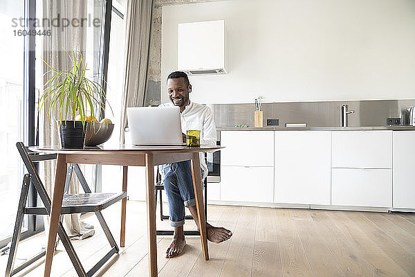 Porträt eines lächelnden Mannes  der an einem Tisch in einer modernen Wohnung sitzt und einen Laptop und Kopfhörer benutzt