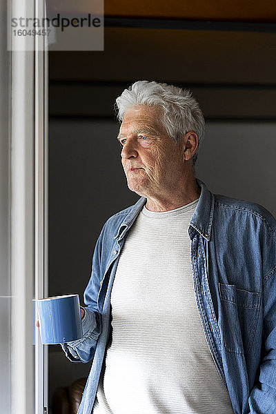 Nachdenklicher älterer Mann  der eine Kaffeetasse hält  während er durch ein Fenster zu Hause schaut