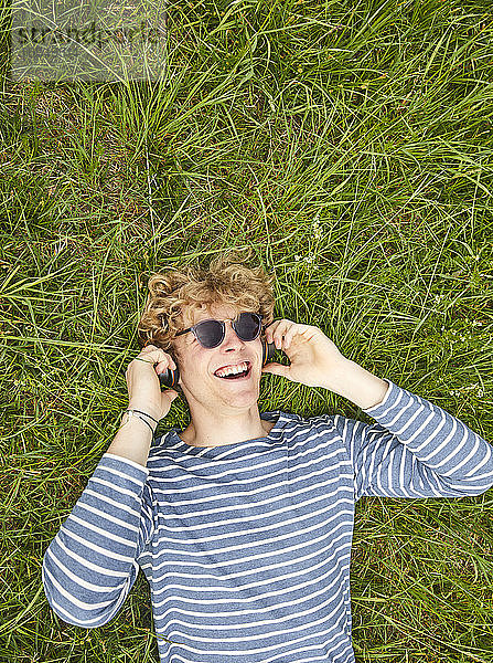 Porträt eines lachenden jungen Mannes mit lockigem blondem Haar  der auf einer Wiese liegt und mit Kopfhörern Musik hört