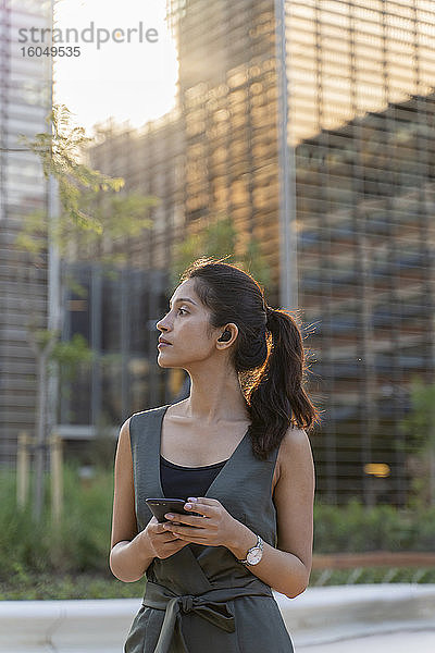 Nachdenkliche Geschäftsfrau hält ihr Smartphone in der Hand  während sie vor modernen Gebäuden in der Stadt steht
