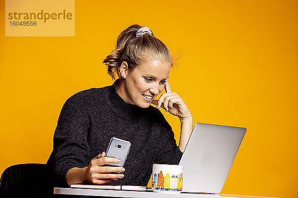 Glückliche schöne Frau  die auf einen Laptop schaut  während sie vor einem gelben Hintergrund sitzt