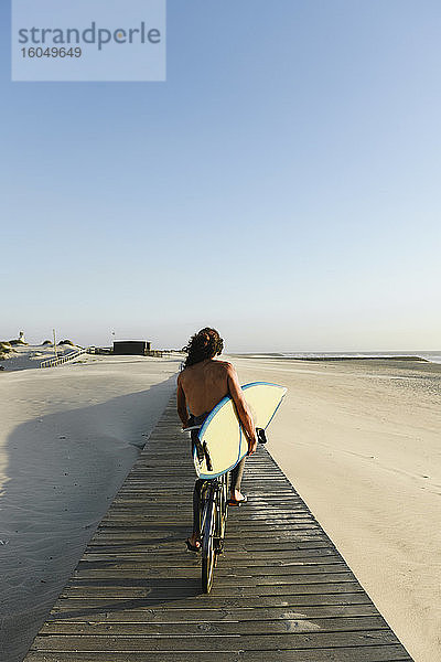 Surfer  der bei Sonnenuntergang Fahrrad fährt und ein Surfbrett hält