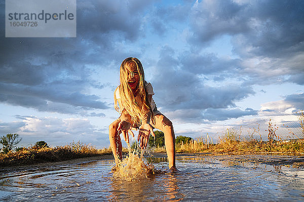 Fröhliches blondes Mädchen spielt beim Spritzen von Wasser auf Pfütze gegen Himmel während Monsun