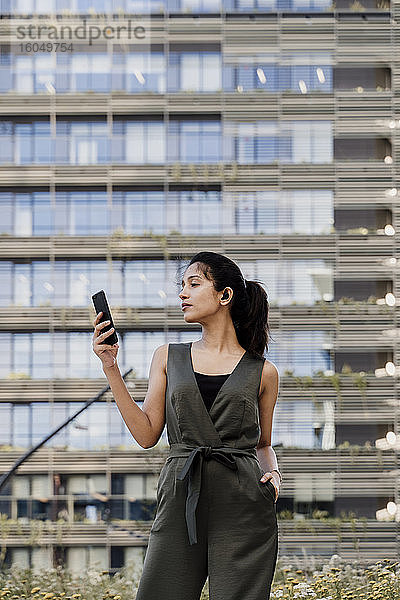 Selbstbewusste Geschäftsfrau  die ein Smartphone benutzt  während sie an einem Gebäude in der Stadt steht