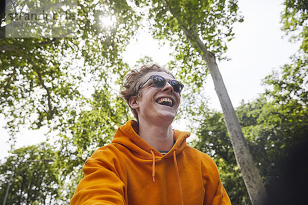 Porträt eines lachenden jungen Mannes mit Sonnenbrille und orangefarbenem Kapuzenshirt in der Natur