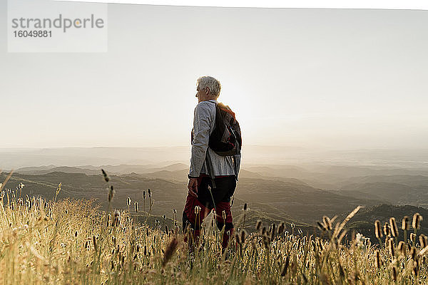 Aktiver älterer männlicher Wanderer steht auf einem Berg und betrachtet die Landschaft bei Sonnenuntergang