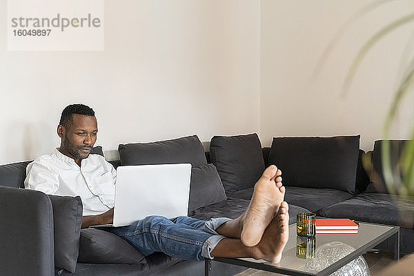 Porträt eines Mannes  der mit hochgelegten Füßen auf der Couch sitzt und einen Laptop benutzt