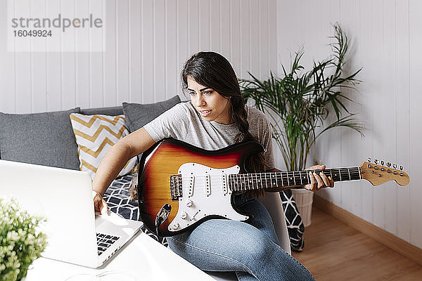 Junge Frau benutzt einen Laptop  während sie zu Hause E-Gitarre spielt