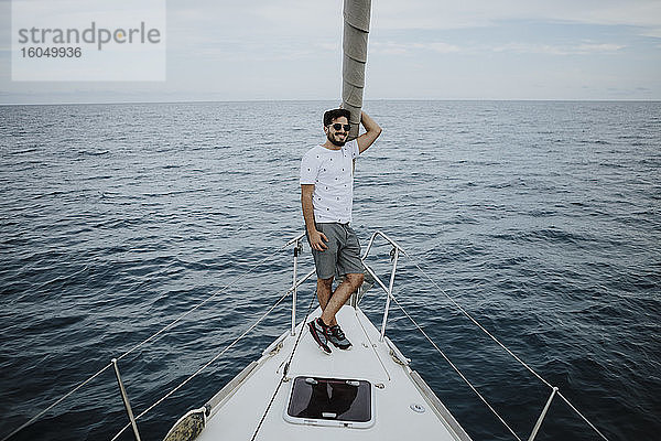 Männlicher Segler mit Sonnenbrille auf dem Bug eines Segelboots im Meer