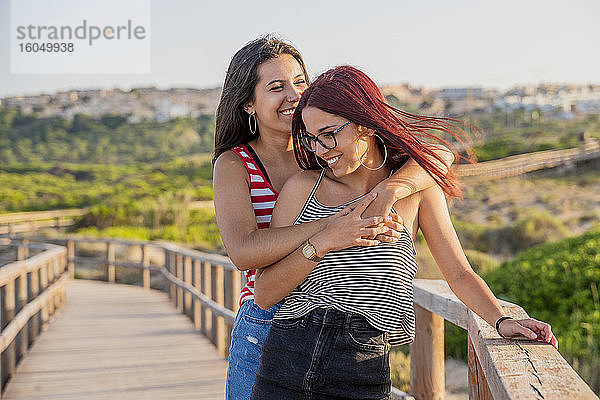 Fröhliche Teenager-Mädchen umarmen sich an der Strandpromenade gegen den klaren Himmel