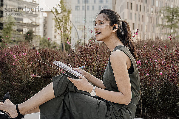 Nachdenkliche Geschäftsfrau mit Notizblock und Mobiltelefon bei Pflanzen in der Stadt sitzend