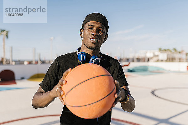 Junger Mann hält Basketball  während er an einem sonnigen Tag vor einem klaren Himmel steht