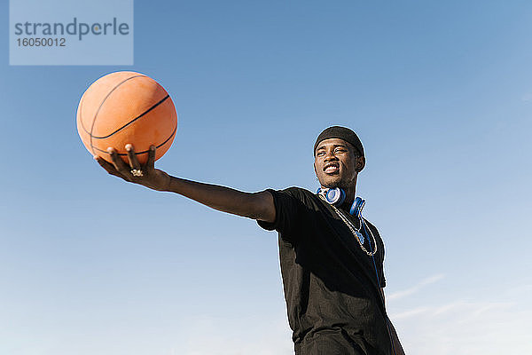 Junger Mann  der einen Basketball hält  während er an einem sonnigen Tag vor einem klaren blauen Himmel steht