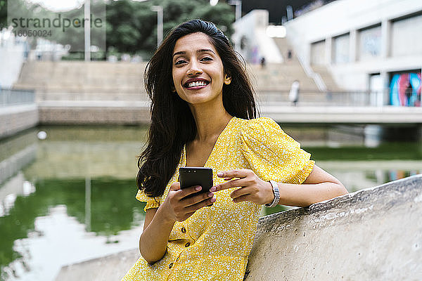 Glückliche junge Frau  die ihr Smartphone in der Hand hält  während sie sich an eine Stützmauer in der Stadt lehnt