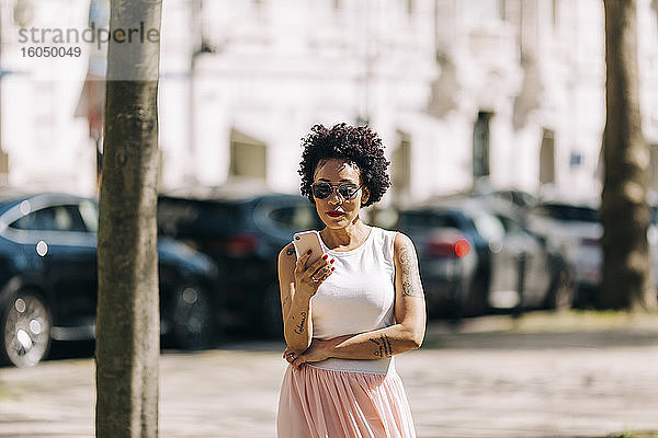 Stilvolle Frau mit lockigem Haar  die in der Stadt stehend ihr Smartphone benutzt