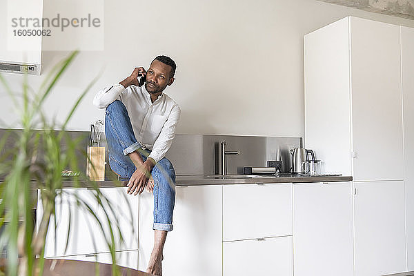 Porträt eines ernsten Mannes am Telefon  der zu Hause auf dem Küchentisch sitzt und in die Ferne schaut