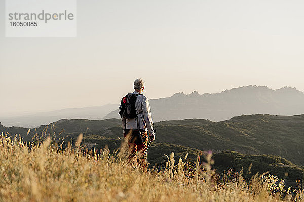 Aktiver älterer Mann mit Rucksack  der bei Sonnenuntergang auf eine Bergkette blickt