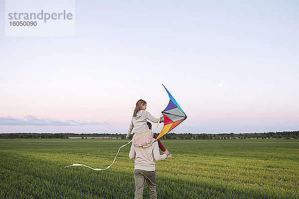 Vater  der seine Tochter auf den Schultern trägt  während er bei Sonnenuntergang über eine Graslandschaft läuft