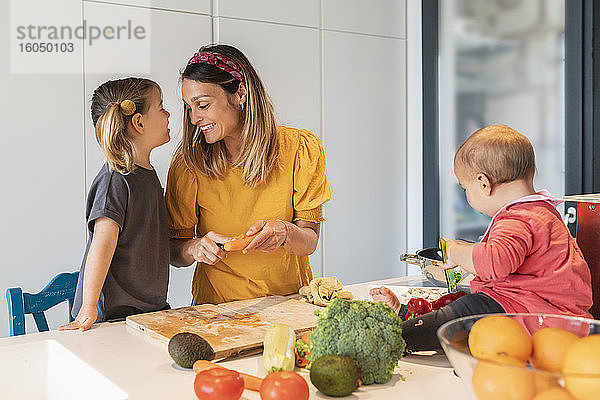 Lächelnde Mutter und Mädchen bereiten Essen zu  während ihre kleine Tochter auf der Kücheninsel spielt