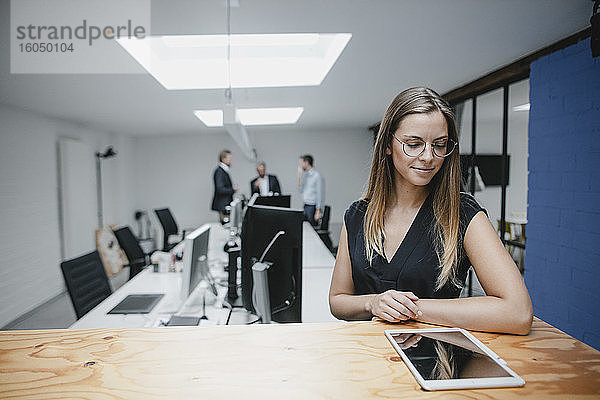 Geschäftsfrau in einem Büro stehend  auf einen Holztisch gestützt  mit einem digitalen Tablet