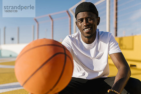 Lächelnder junger Mann  der einen Basketball hält  während er an einem sonnigen Tag im Gericht sitzt