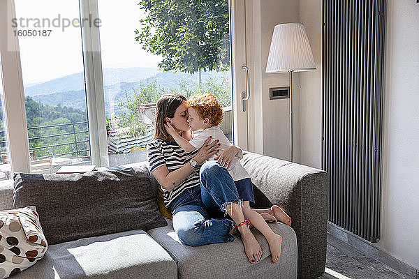 Mutter küsst süßen Sohn  während sie auf dem Sofa im Wohnzimmer zu Hause sitzt