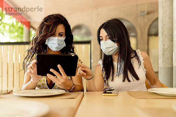 Junge Frauen  die während des Coronavirus in einem Restaurant sitzen und sich ein digitales Tablet teilen