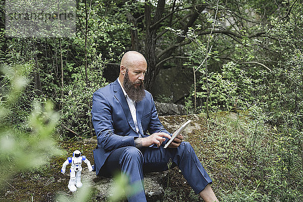 Glatzköpfiger Geschäftsmann  der ein digitales Tablet benutzt und mit einem Roboter inmitten von Bäumen im Wald sitzt