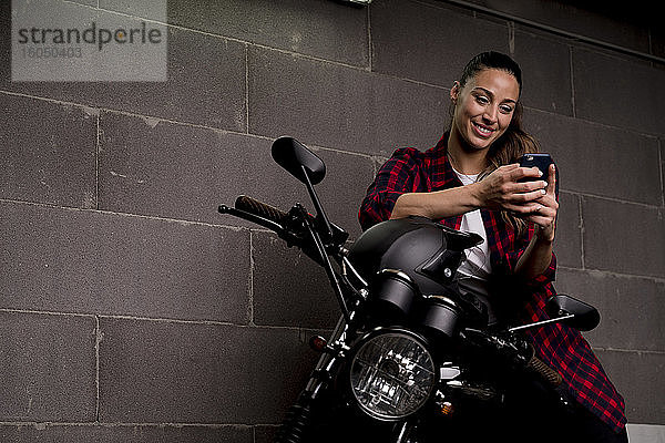 Junge lächelnde Frau mit Smartphone auf dem Motorrad sitzend