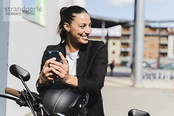 Lächelnde Frau  die auf einem Motorrad sitzt und ein Smartphone benutzt