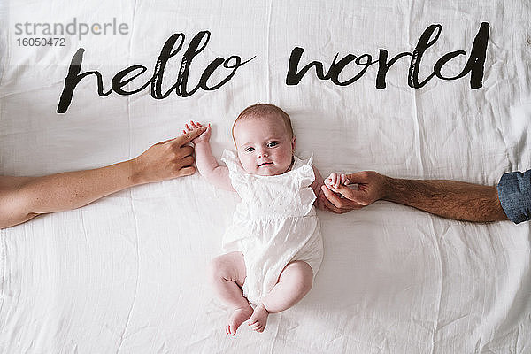 Happy cute baby girl holding hands of parents while lying on blanket under Hello World text at home