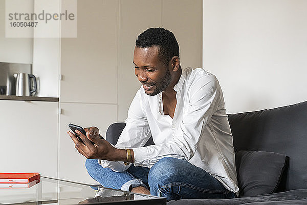 Lächelnder Mann sitzt zu Hause auf der Couch und benutzt sein Smartphone