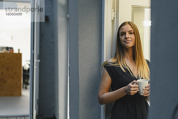 Geschäftsfrau steht in offener Bürotür und hält eine Tasse Kaffee